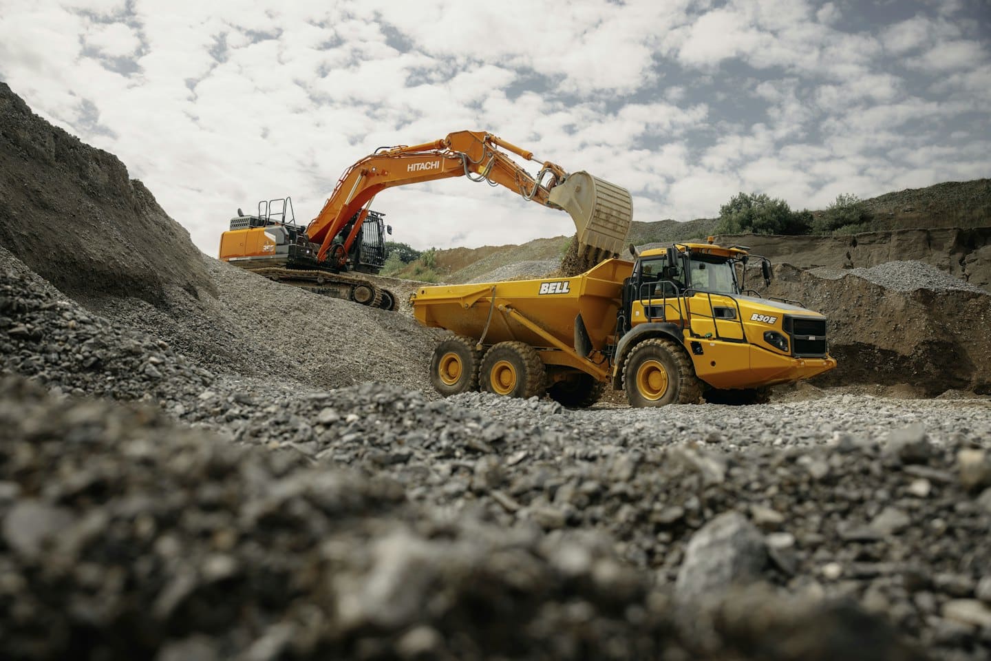 Orange excavator fills yellow dump truck with gravel.